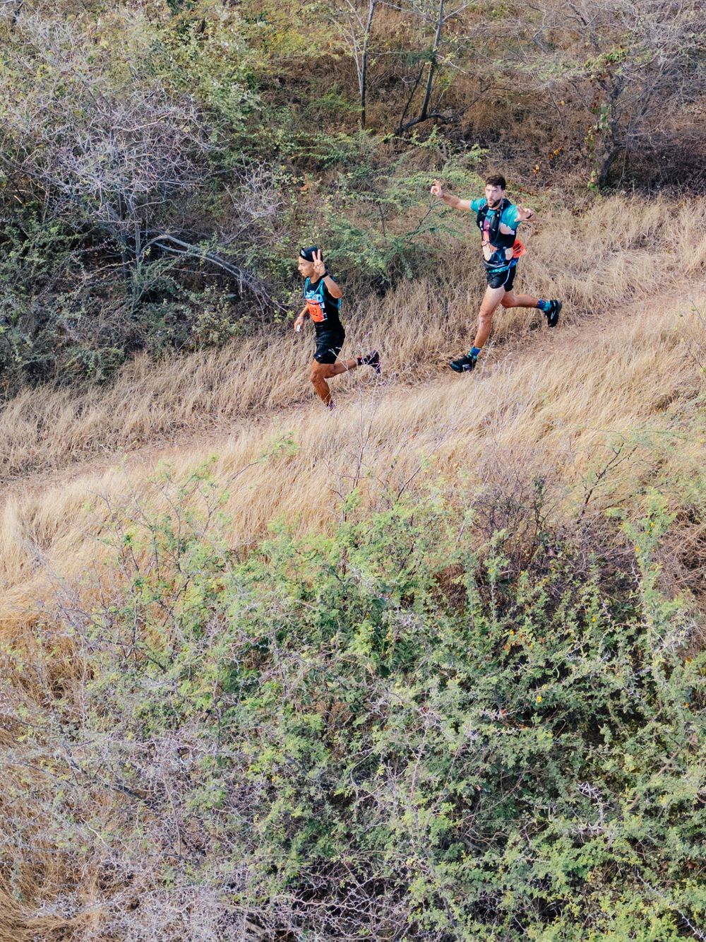 Two trail runners navigate a steep hillside trail through dry grass and brush during an outdoor workout session.