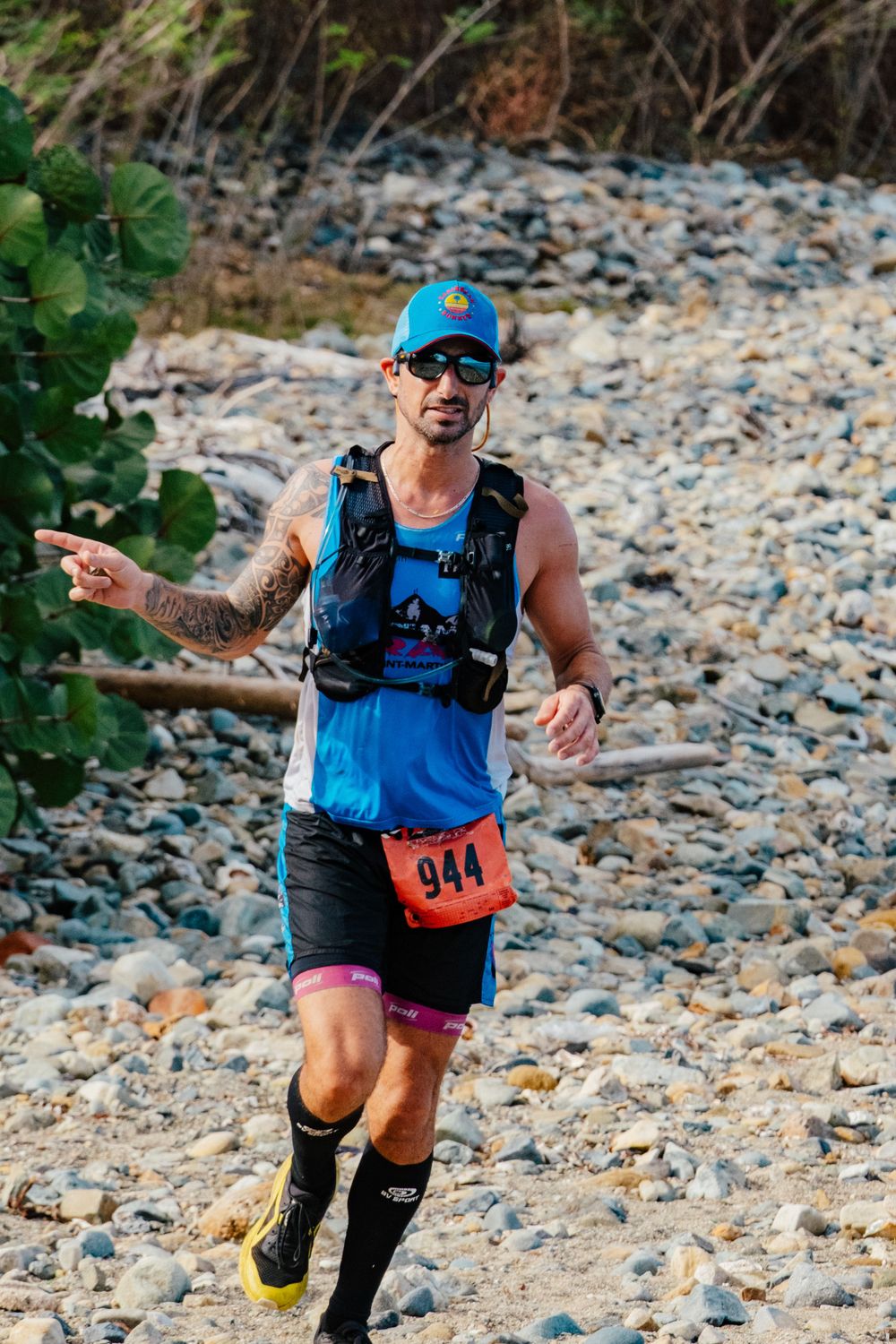 Trail runner wearing blue tank top and hydration pack competes on rocky terrain during endurance race.