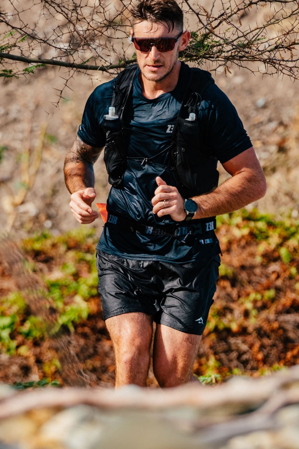 Trail runner in black gear and sunglasses moving through rugged outdoor terrain during competition.