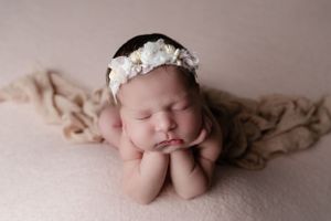 Sleeping newborn lying on pink blanket wearing white lace headband in frog pose during professional photo session.