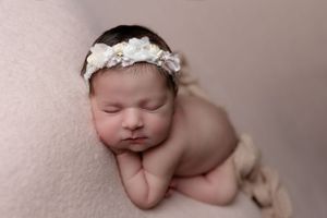 Sleeping newborn wearing delicate white floral headband posed on soft pink backdrop in professional studio setting.