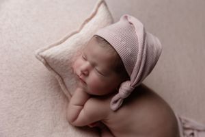 A sleeping newborn baby wearing a pink knit bonnet rests peacefully on a soft pillow.