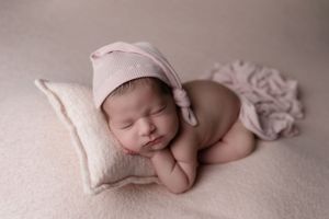 Sleeping newborn wearing soft pink cap rests on cream pillow in artistic pose.
