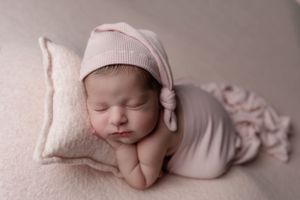 A sleeping newborn in pink attire rests peacefully on a soft beige pillow.