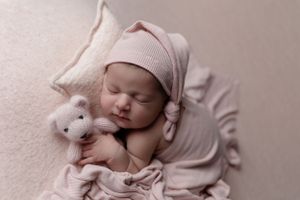 Sleeping newborn wearing pink bonnet cuddling small plush bunny toy on white blanket during professional photo session.