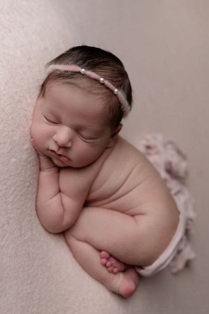 Sleeping newborn wearing delicate headband rests in curled position on soft beige background.