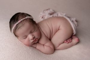 Peaceful sleeping newborn baby wearing delicate lace headband rests on soft pink blanket in curled position.
