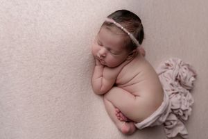 Newborn baby sleeping peacefully in curled position wearing white diaper and delicate headband on soft pink backdrop.