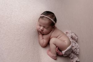 Peaceful newborn baby wearing headband resting on soft pink background.