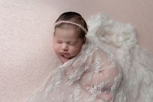 Sleeping newborn draped in delicate white lace with pearl headband on pink backdrop.