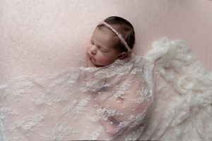 Newborn baby covered in ethereal white lace rests peacefully on pink background.