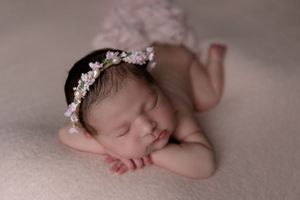 Sleeping newborn with floral headpiece rests on beige blanket in artistic photography pose.