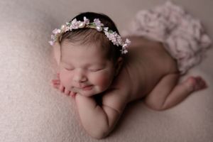 Sleeping newborn wearing floral crown poses on soft pink background in artistic portrait.