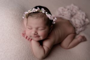 Newborn wearing floral crown lies peacefully on blush pink backdrop during professional photo session.