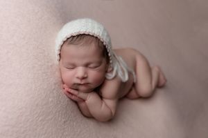 Peaceful newborn baby wearing white knit bonnet poses on pink background.