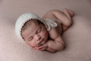 Newborn baby sleeping peacefully on pink fabric wearing a white knitted bonnet.