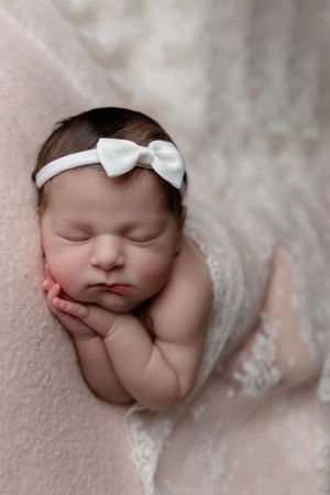 Sleeping newborn baby wrapped in pink lace wearing white bow headband while resting peacefully.