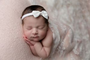 Peaceful sleeping newborn baby wrapped in white lace blanket with bow headband on pink backdrop.