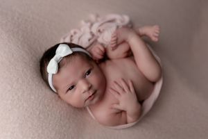 Tiny newborn lying on pink fabric wearing white bow headband.