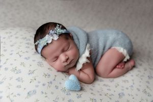 Peaceful newborn baby sleeping on floral fabric wearing grey onesie and delicate headband with small blue toy.