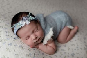 Peaceful newborn sleeping in gray wrap wearing floral headpiece on textured blanket.