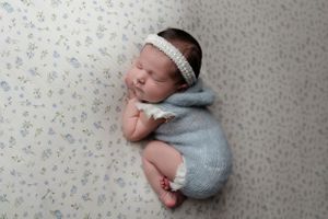 Sleeping newborn in white headband and blue outfit rests on floral patterned fabric.