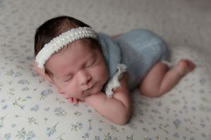 Newborn baby in white headband and blue romper sleeping on floral print backdrop.