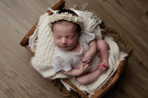 Sleeping newborn wrapped in delicate white lace blanket resting peacefully in wooden basket on hardwood floor.