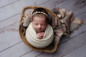 Sleeping newborn in cream knit wrap nestled in woven basket on wooden floor.