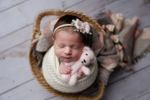 Peaceful newborn in white knit wrap sleeping in basket with small teddy bear and floral headband.