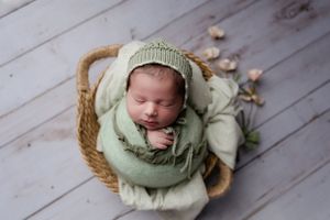 Newborn baby in mint green wrap sleeping in woven basket on wooden floor.