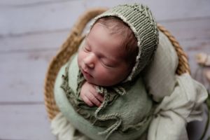 Newborn baby sleeping in sage green knit bonnet and wrap in wooden basket.