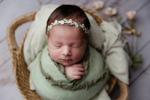 Sleeping newborn wrapped in mint green swaddle wearing delicate headband resting in woven basket.