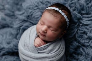 Sleeping newborn swaddled in gray wrap with delicate floral headband on blue textured backdrop.