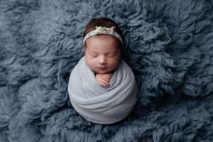 Swaddled newborn wearing delicate crown rests on textured blue-grey backdrop.