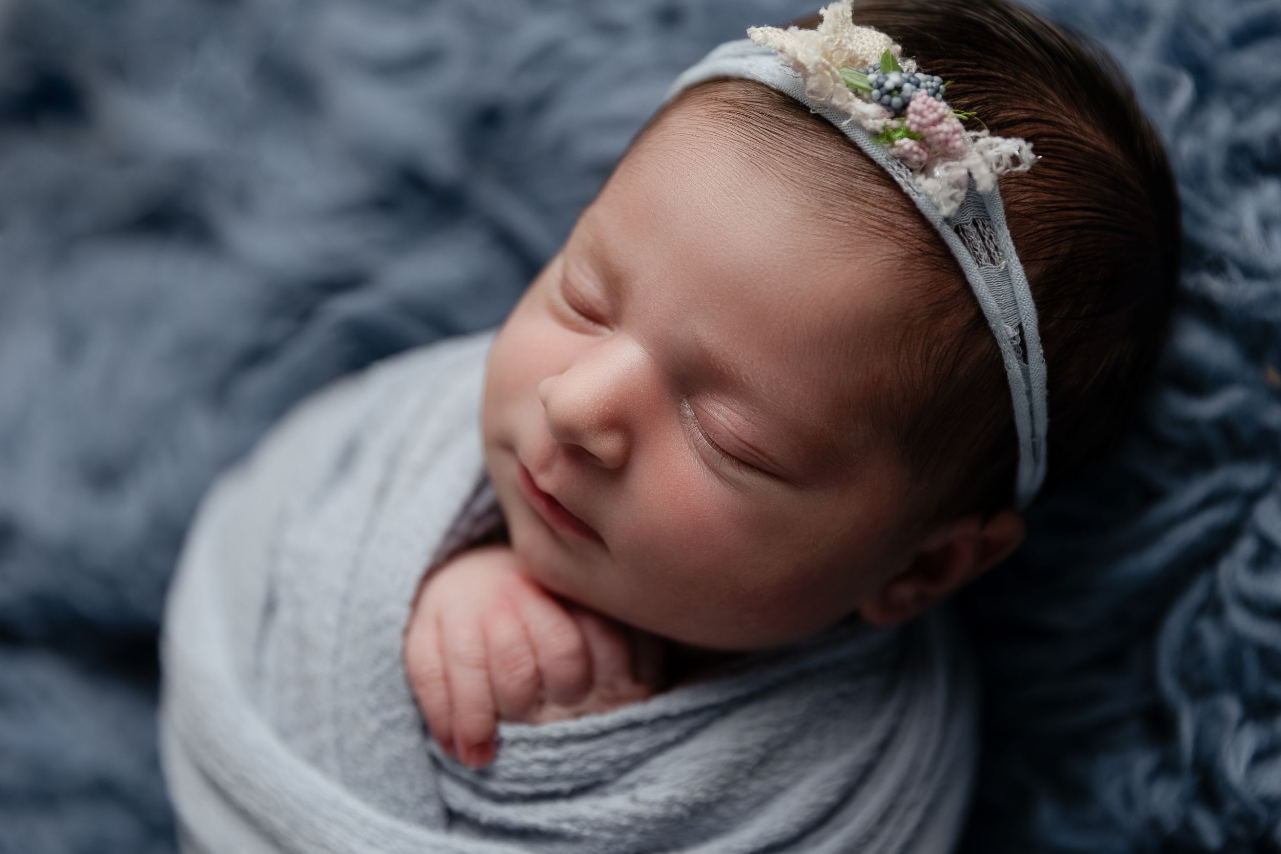 Peaceful newborn wrapped in soft gray swaddle with lace headband against blue textured backdrop.