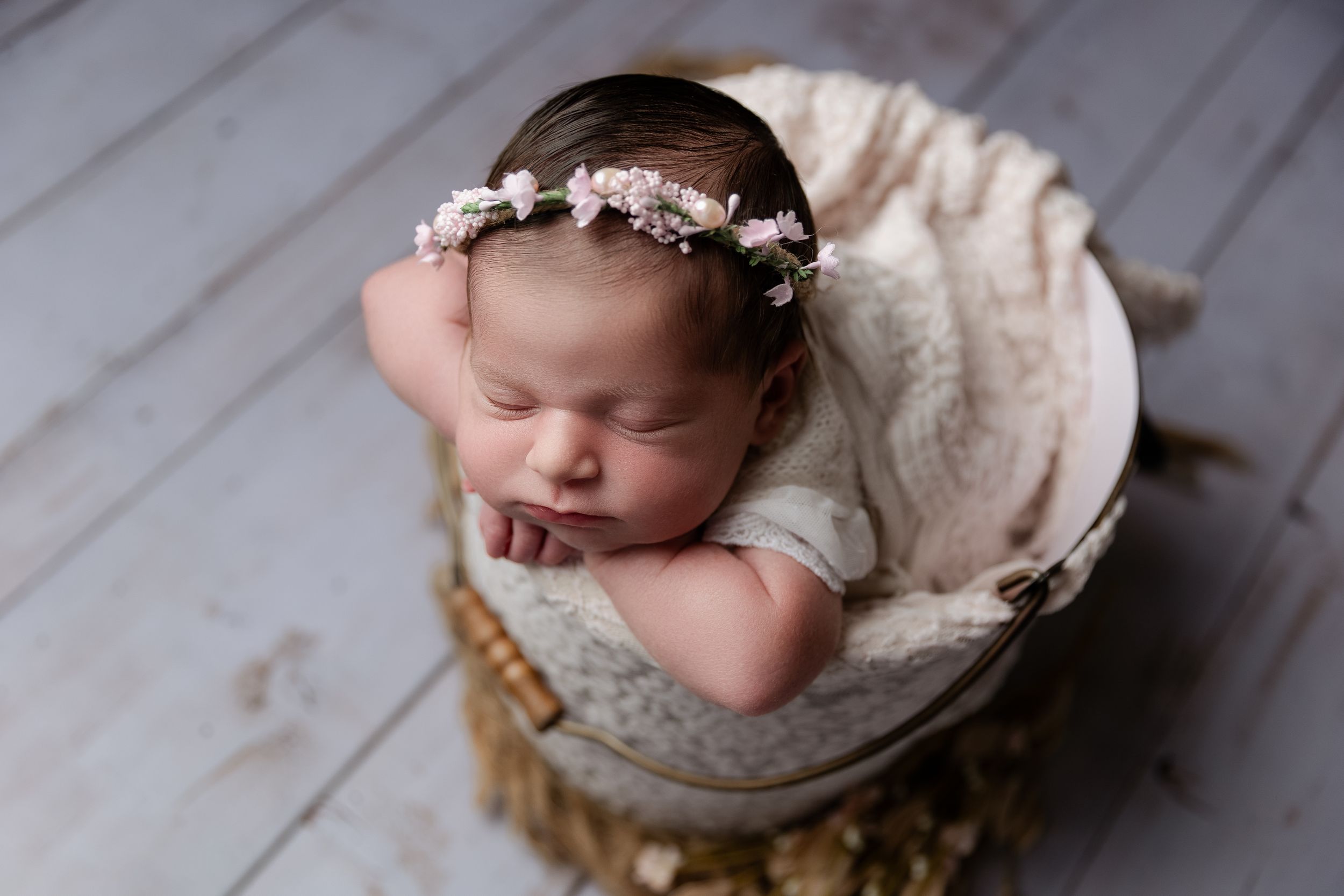 Sleeping newborn in cream outfit rests in wooden basket wearing delicate floral headband.