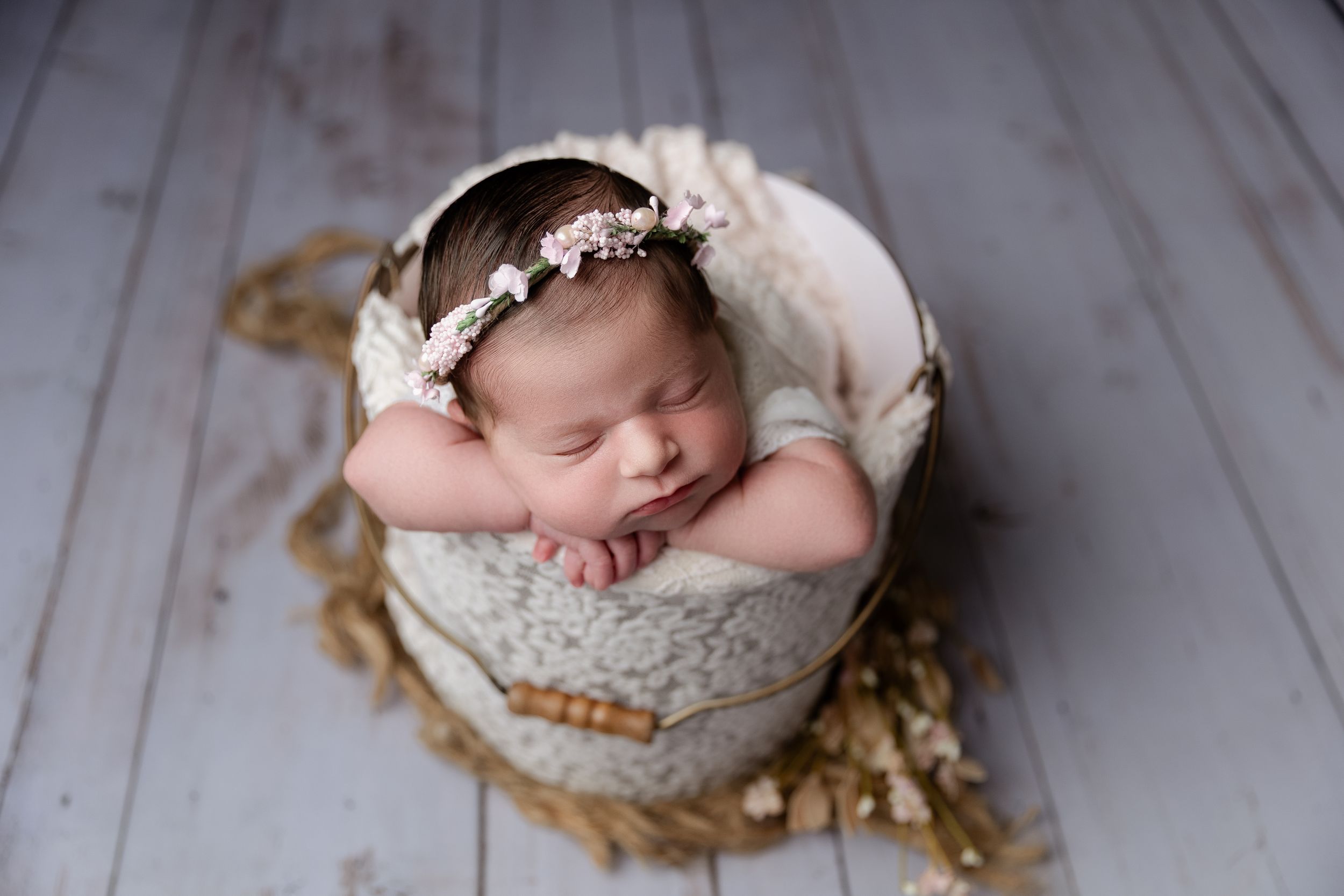 Sleeping newborn baby nestled in cream bowl on wooden floor with delicate floral crown and white fabric.