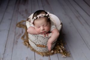 A sleeping newborn rests in a rustic wooden bowl adorned with a delicate white floral headband.