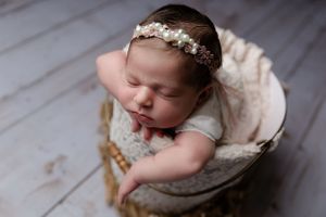 Sleeping infant wearing a delicate floral headband rests peacefully in a cream-colored basket on a wooden floor.