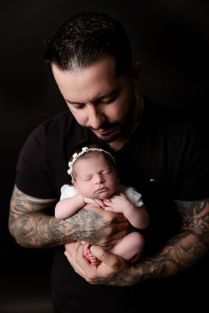An infant in white clothing sleeps peacefully while cradled in tattooed arms against a dark background.