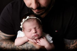 A sleeping newborn baby wearing white clothing and a delicate headband rests peacefully against a dark background.