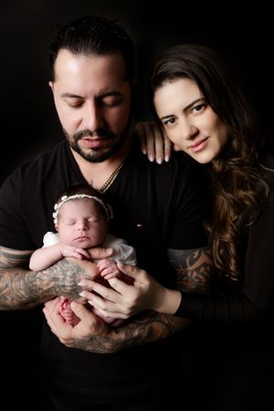 A couple dressed in black holds their sleeping newborn baby against a dark background in an intimate studio portrait.