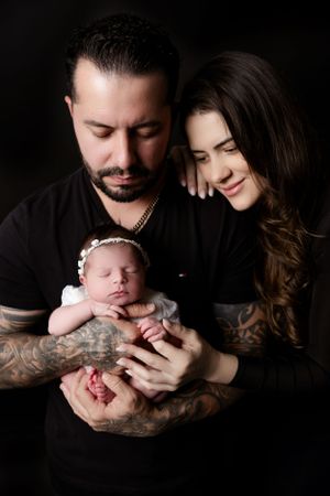 A tender family portrait showing parents holding their sleeping newborn baby against a dark background.
