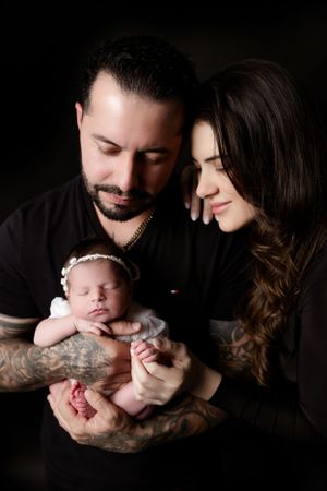 A tender family moment captured in dark tones as parents gaze lovingly at their sleeping newborn baby.
