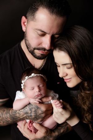 Intimate family portrait featuring a sleeping newborn in white with loving parents against dark background.
