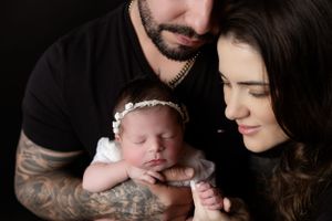 A peaceful newborn baby wearing a white headband is held lovingly by parents in a studio portrait against dark background.