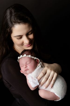 A newborn baby wearing white swaddle and floral headband is held tenderly against a dark background.