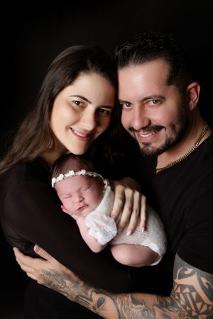 A family portrait against a dark background shows parents holding their newborn baby wrapped in white lace.