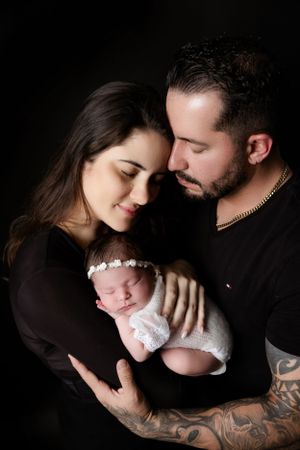 A sleeping newborn in white wrap and floral headband is cradled by parents wearing dark clothes.